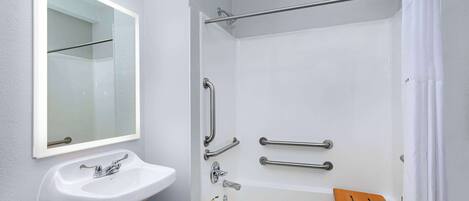 A bathroom with a white sink, a mirror, a shower with grab bars, and a wooden bath mat.