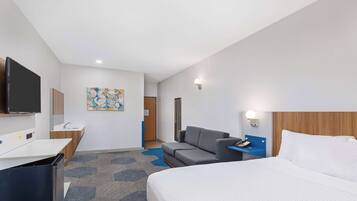Quarto de hotel com uma cama, uma escrivaninha com televisão de tela plana, um sofá, uma mesinha com telefone e uma luminária na parede.
