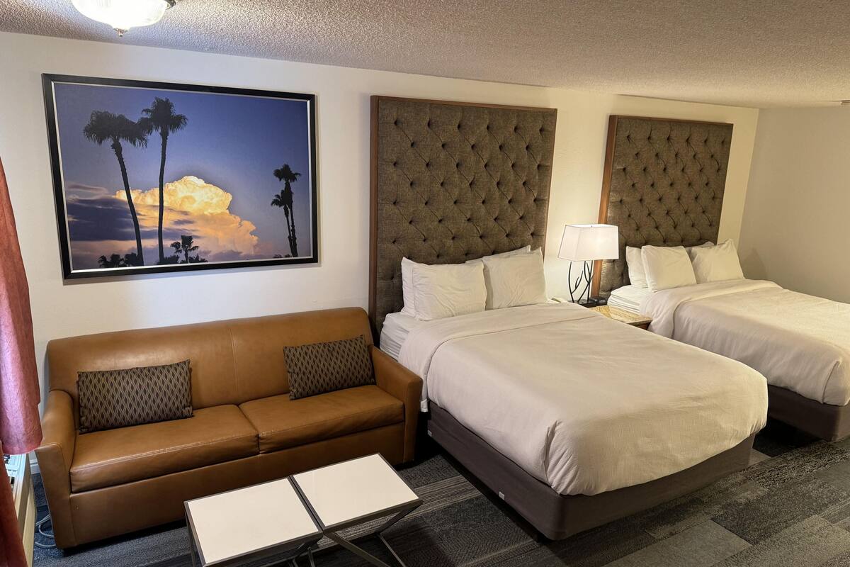 Una habitación de hotel con un sofá de cuero marrón, dos camas con cabeceros capitonados, un cuadro grande enmarcado con palmeras y una mesita.
