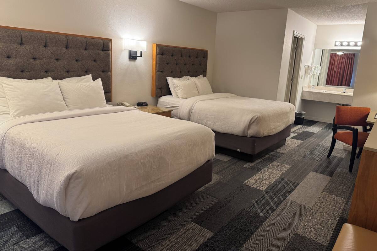 Habitación de hotel con dos camas, una mesita, una silla, una mesita de noche, una lámpara de pared y un ventanal con cortinas.
