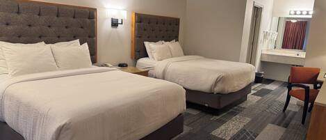 Habitación de hotel con dos camas, una mesita, una silla, una mesita de noche, una lámpara de pared y un ventanal con cortinas.