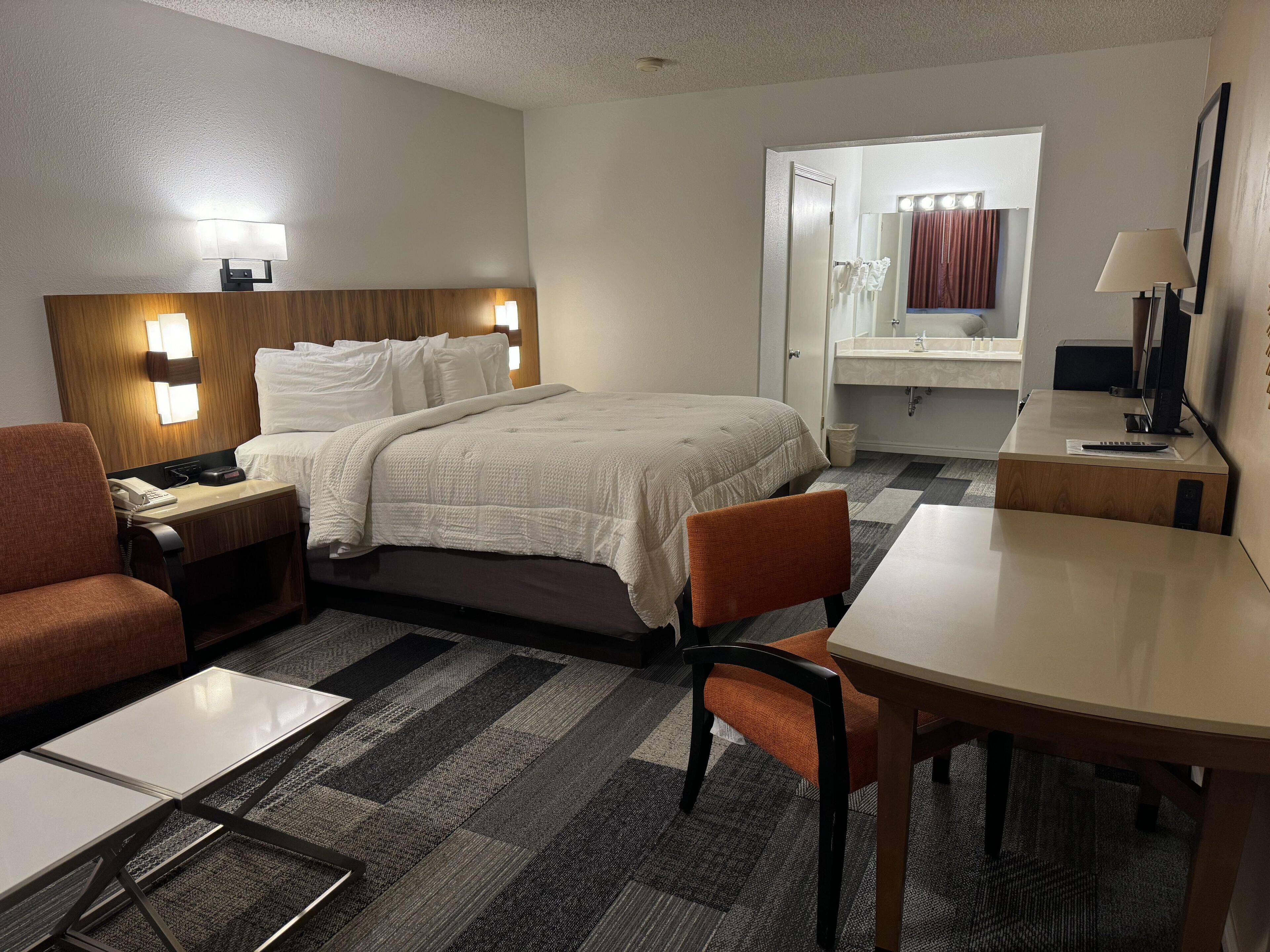A hotel room with a bed, a desk, a chair, a nightstand, a lamp, a mirror, and a bathroom with a sink and mirror.