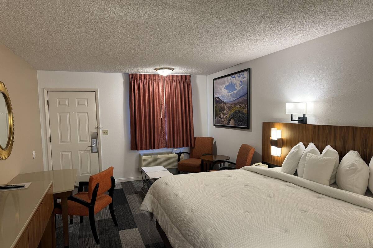 Una habitación de hotel con cama, escritorio, dos sillas, ventana con cortinas, un cuadro en la pared y una lámpara de pared.