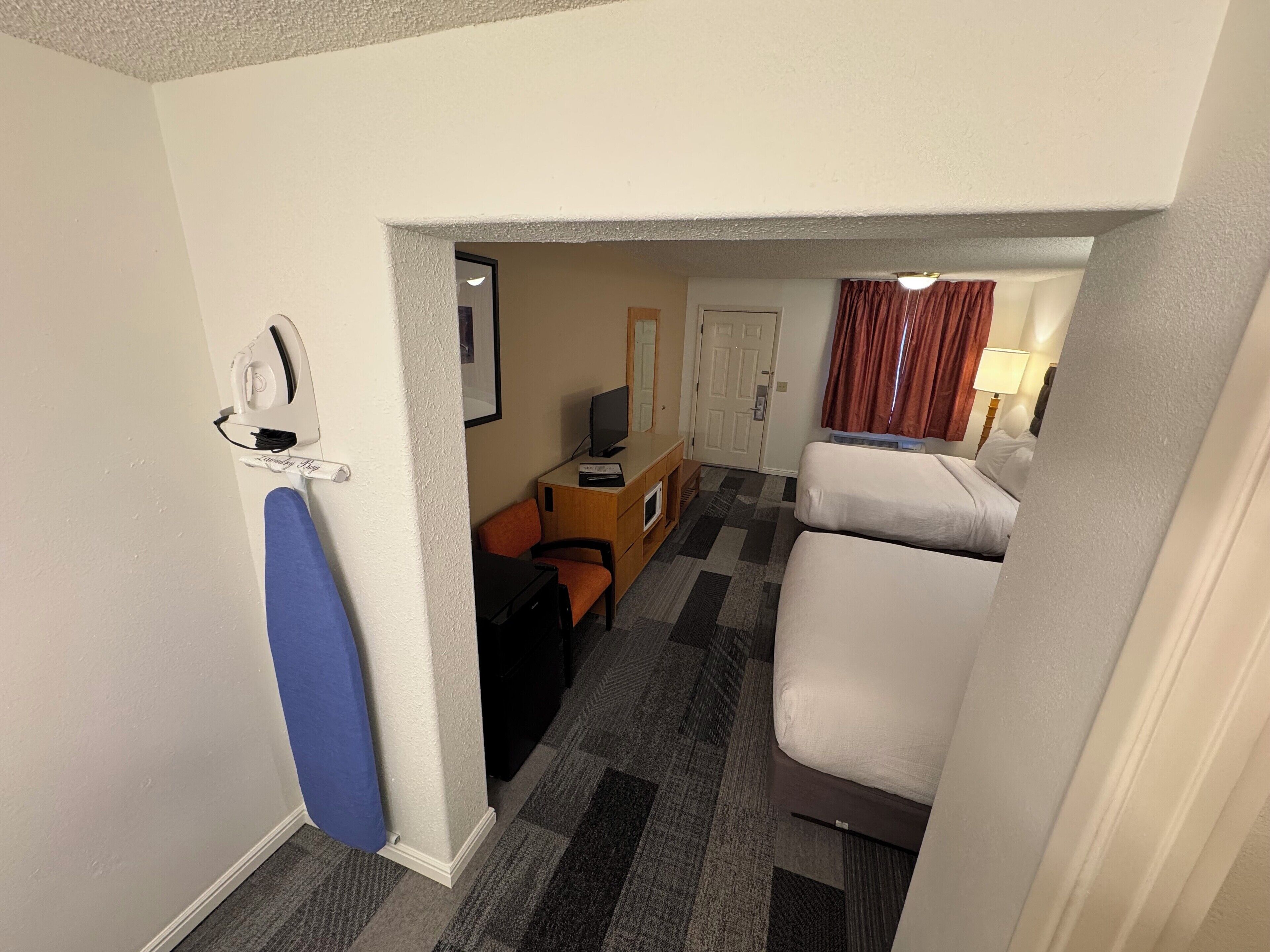 Habitación de hotel con mostrador de recepción, escritorio con computadora, televisor, dos camas y ventana con cortinas.