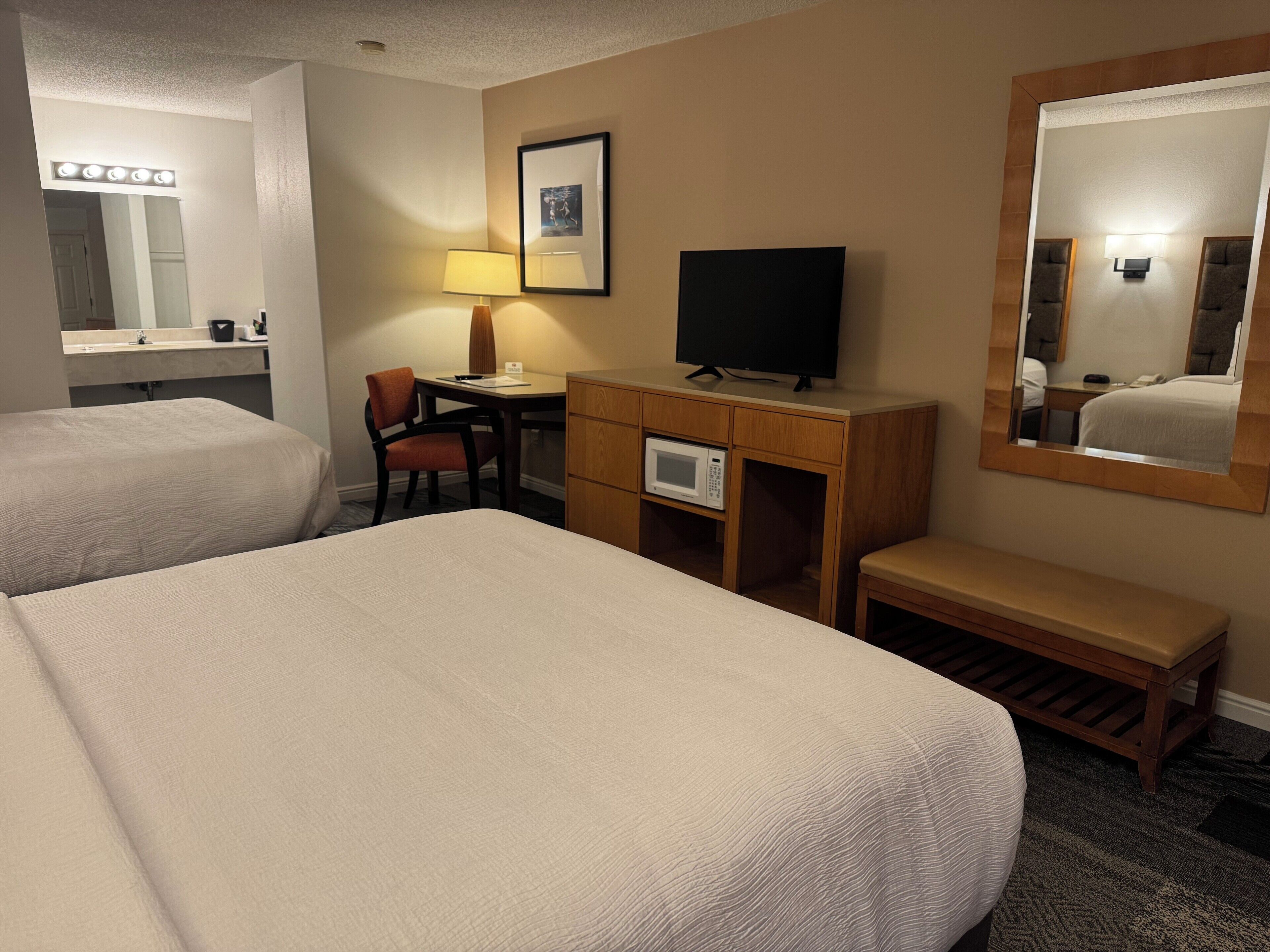 A hotel room with two beds, a desk with a lamp, a television, a microwave, and a large mirror.