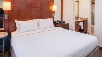 A hotel room with a large bed, a wooden headboard, a bedside table with a lamp, a bathroom with a shower curtain, and a mirror.