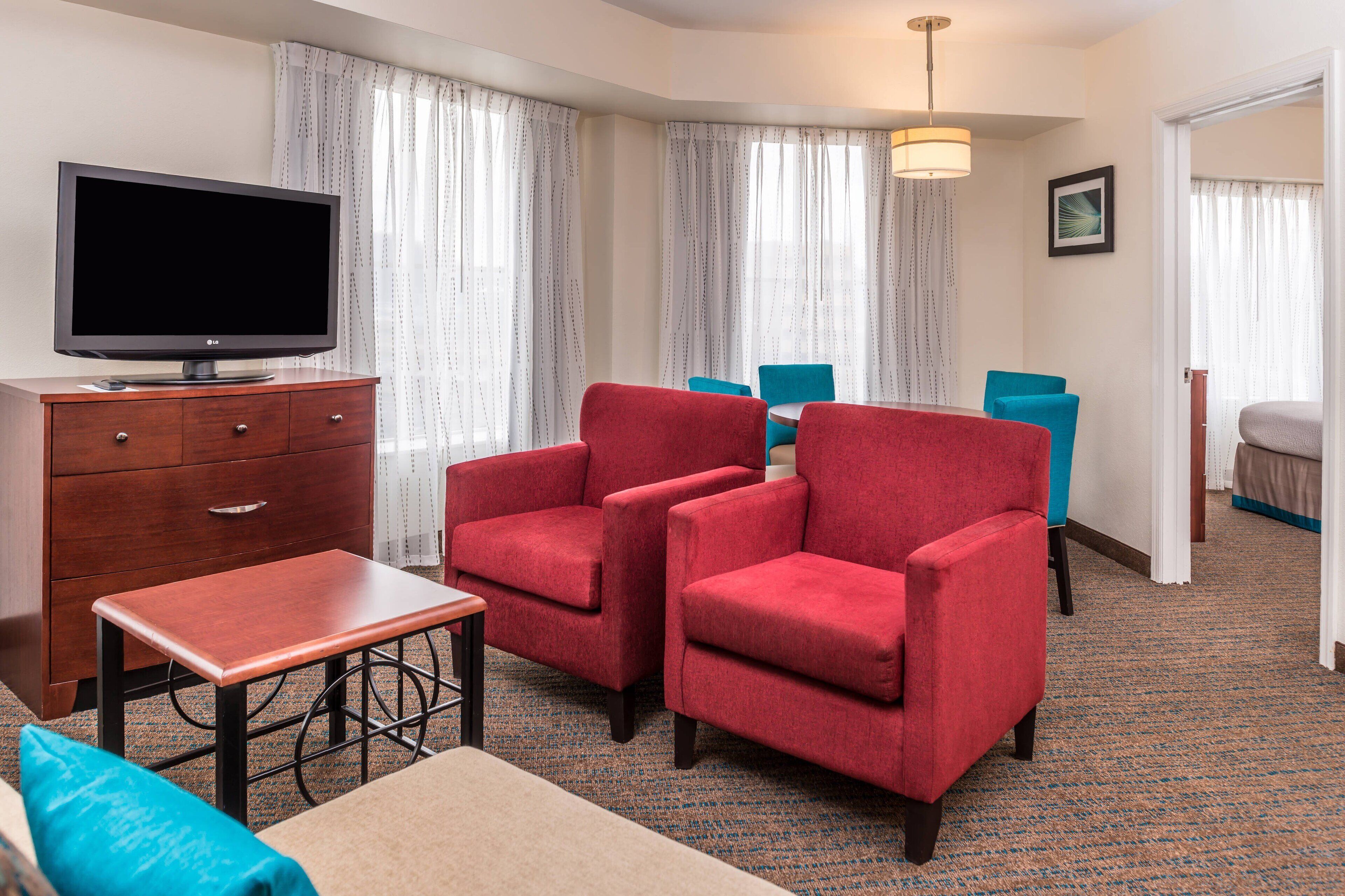 Une chambre d’hôtel équipée d’un téléviseur à écran plat, d’une commode en bois, d’une petite table, de deux fauteuils rouges, d’un fauteuil bleu et d’un lit recouvert d’une couvre-lit bleue et blanche.