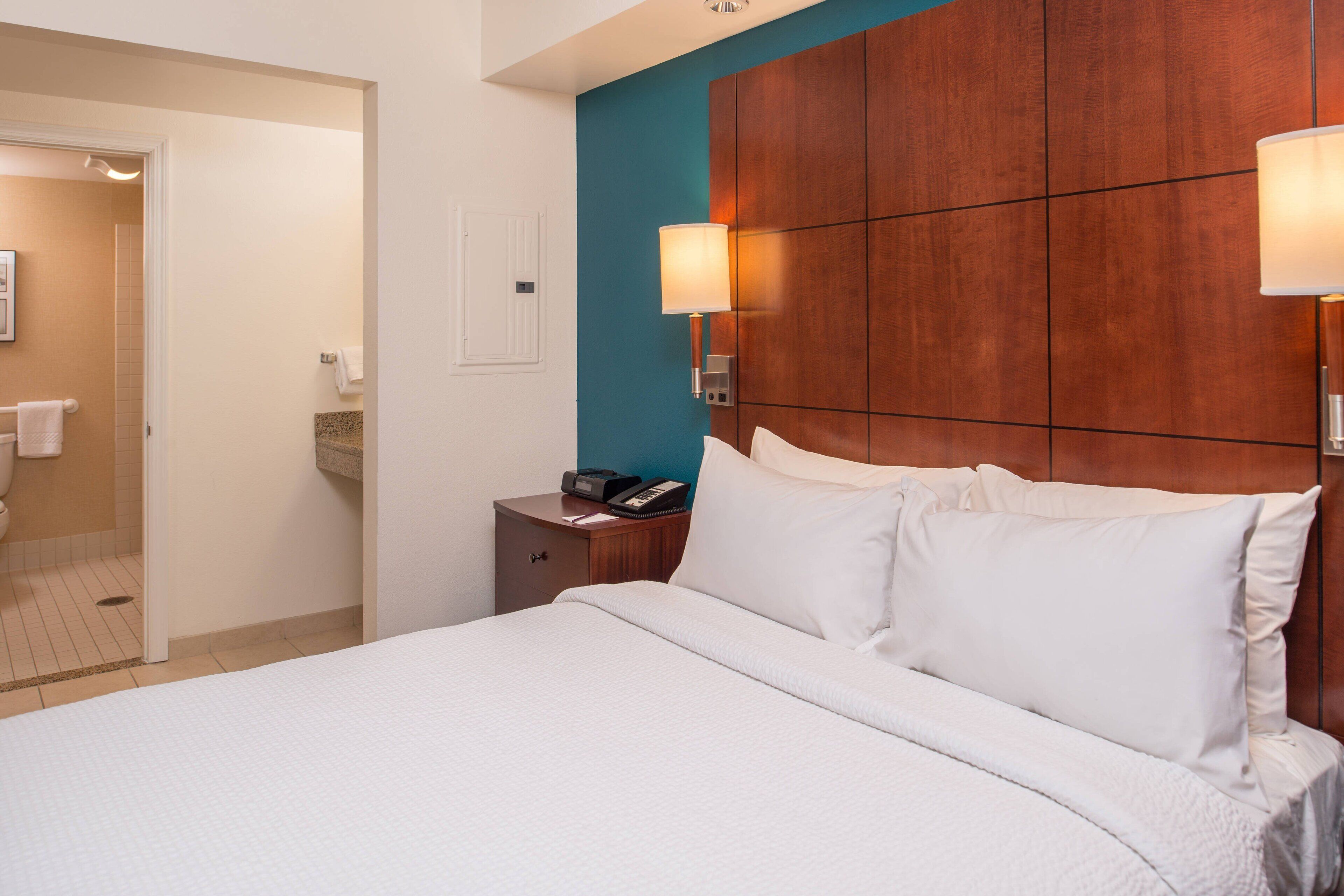 A hotel room with a large bed, a wooden headboard, a nightstand with a phone, and a bathroom visible through an open door.