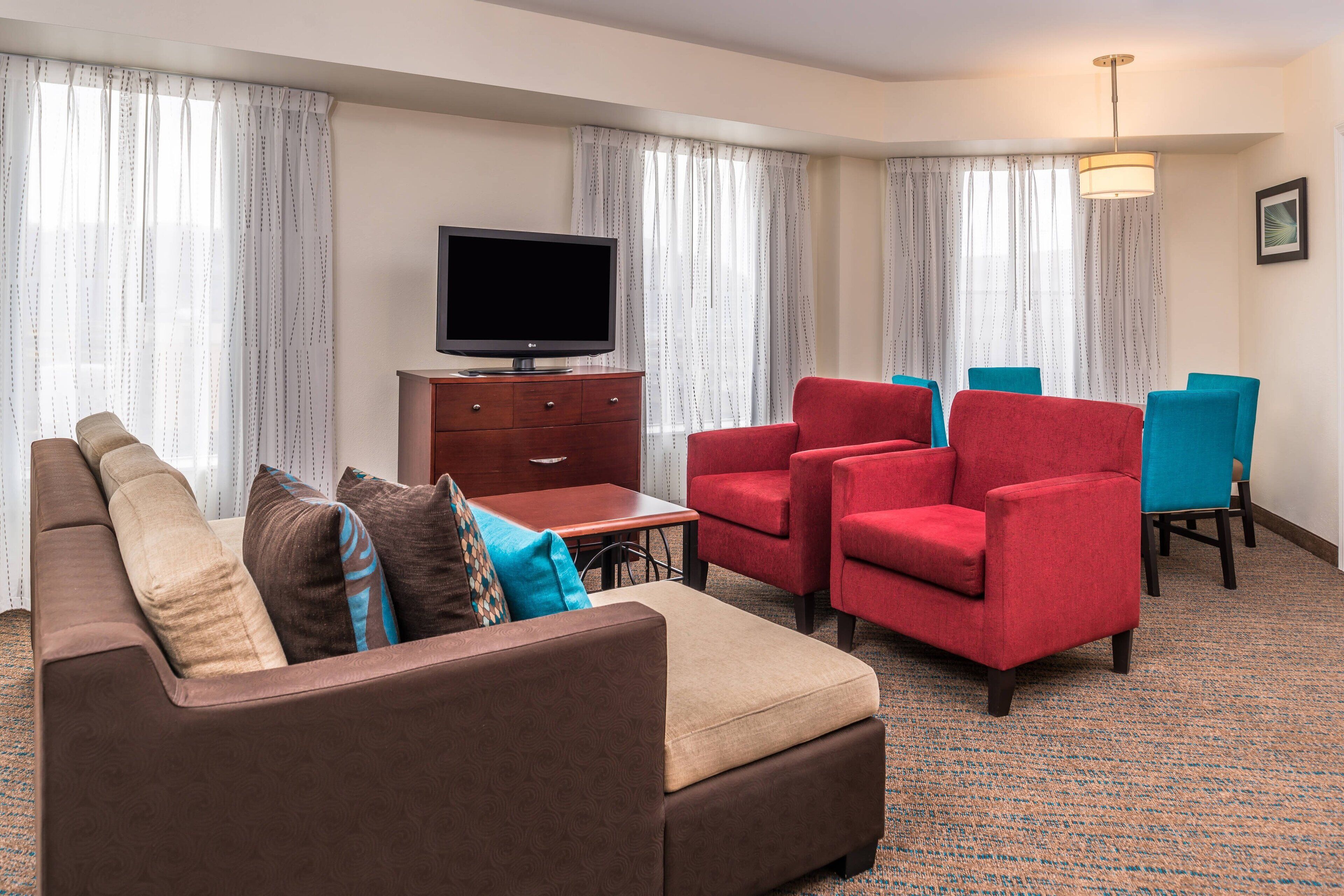 Habitación de hotel con un sofá marrón, sillones rojos, una mesa de madera, un televisor sobre un mueble bajo y una alfombra estampada.