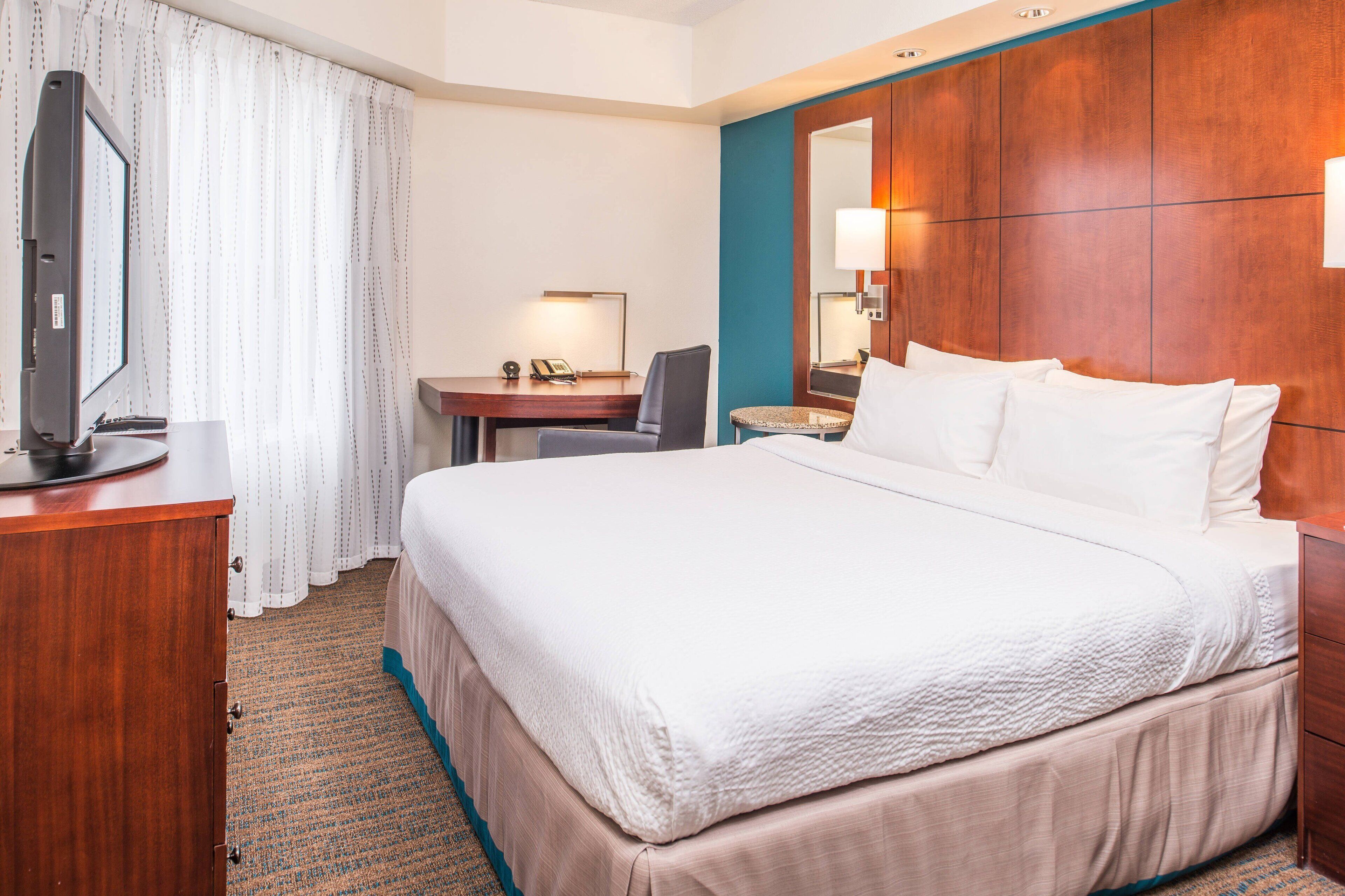A hotel room with a bed, a desk, a chair, a television, and a bathroom mirror.