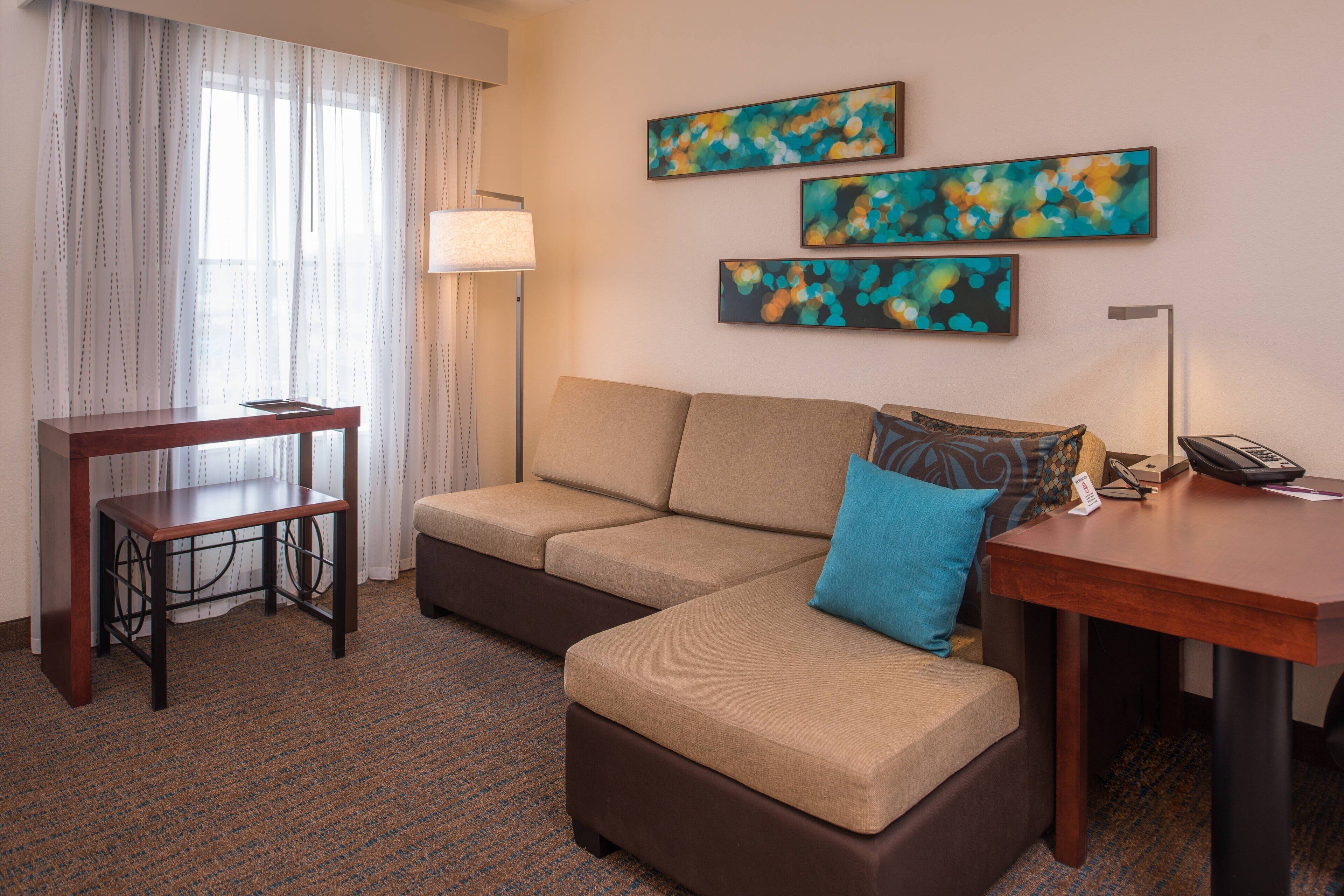 A hotel room with a sofa, a small table, a desk, and a lamp. There are three abstract paintings on the wall.