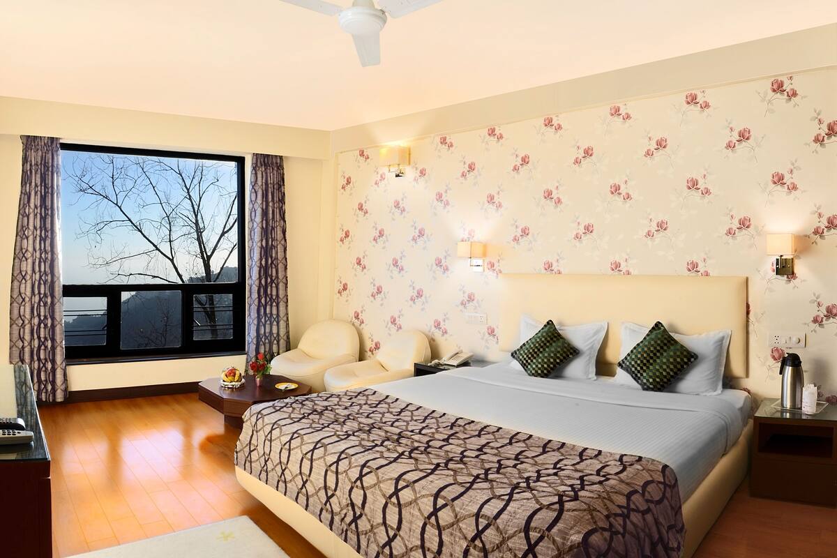 A hotel room with a large bed, two bedside tables, a nightstand, a window with curtains, a ceiling fan, and a floral wallpaper.