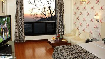 A hotel room with a large window, a bed, a sofa, a television, and a small table with a vase of flowers.