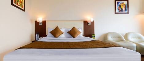 A hotel room with a large bed, two wall-mounted lamps, two paintings, and a pair of white armchairs.