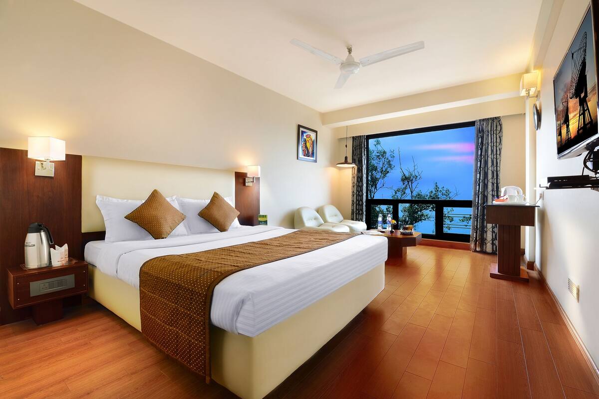 A hotel room with a large bed, a desk, a chair, a TV, and a balcony with a view.