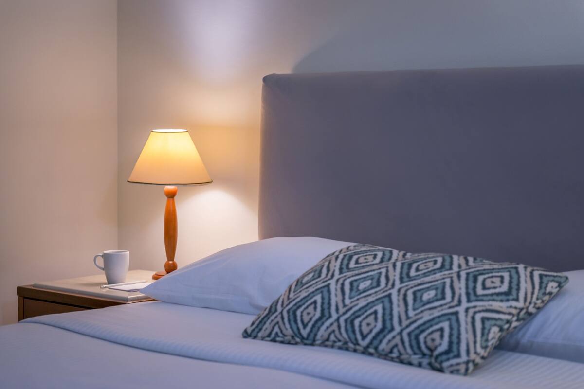 Een net opgemaakt bed met een kussen met patroon, een nachtkastje met een lamp en een beker.