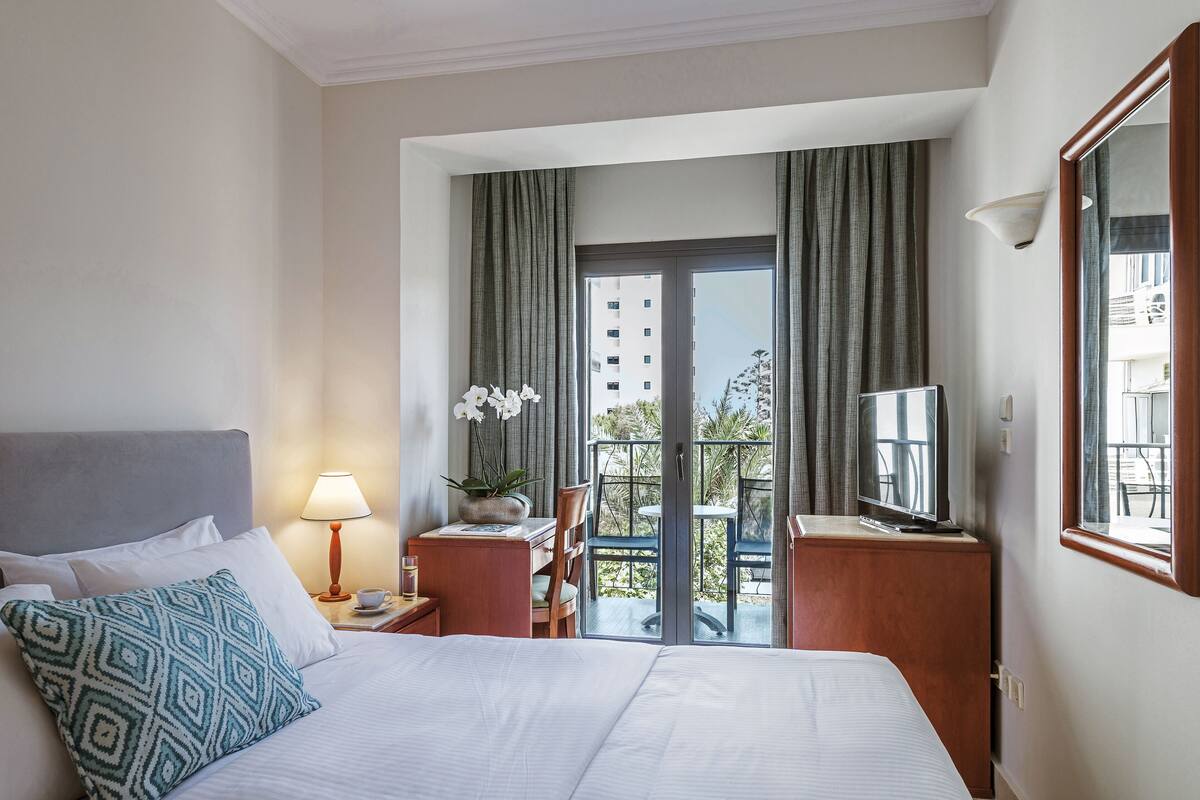Een hotelkamer met een bed, een bureau, een stoel, een lamp, een televisie en een balkon met gordijnen.