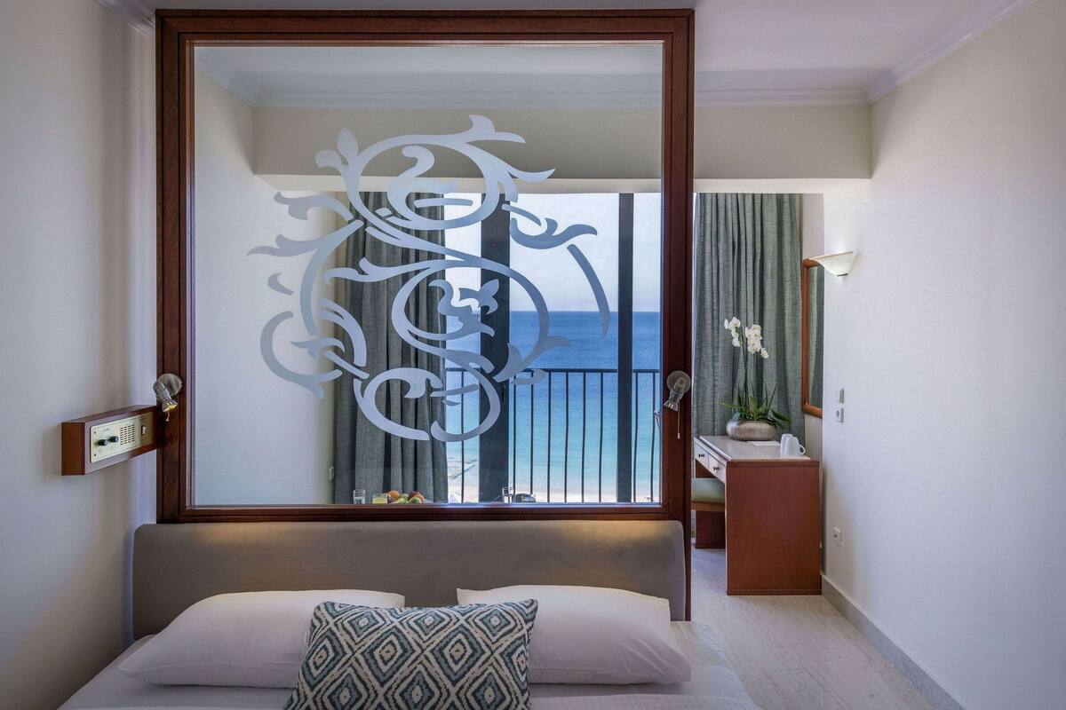 Een hotelkamer met een bed, een decoratieve spiegel, uitzicht op zee en een klein tafeltje met een plant.