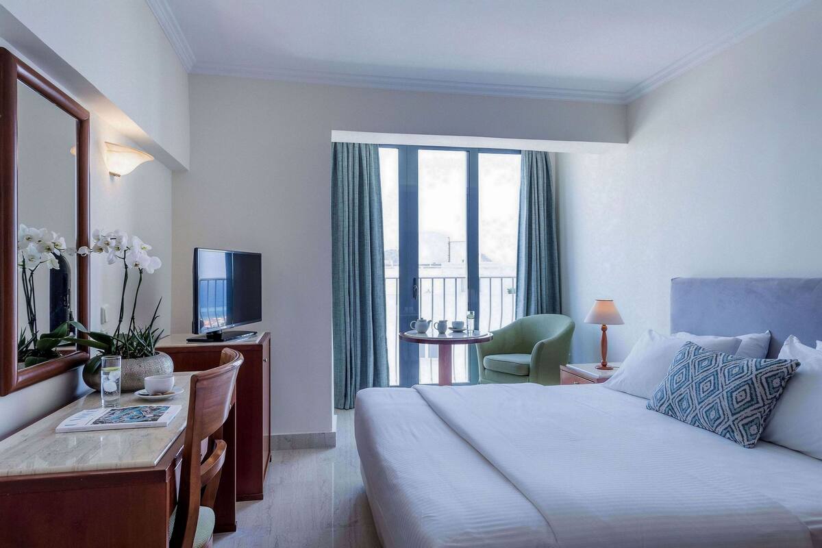 Een hotelkamer met een bed, een bureau met stoel, een televisie, een tafeltje met een lamp en een balkon met gordijnen.