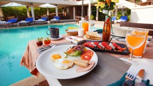Un desayuno junto a la piscina, con un plato de huevos, jamón y verduras, un vaso de zumo de naranja y un arreglo floral tropical.