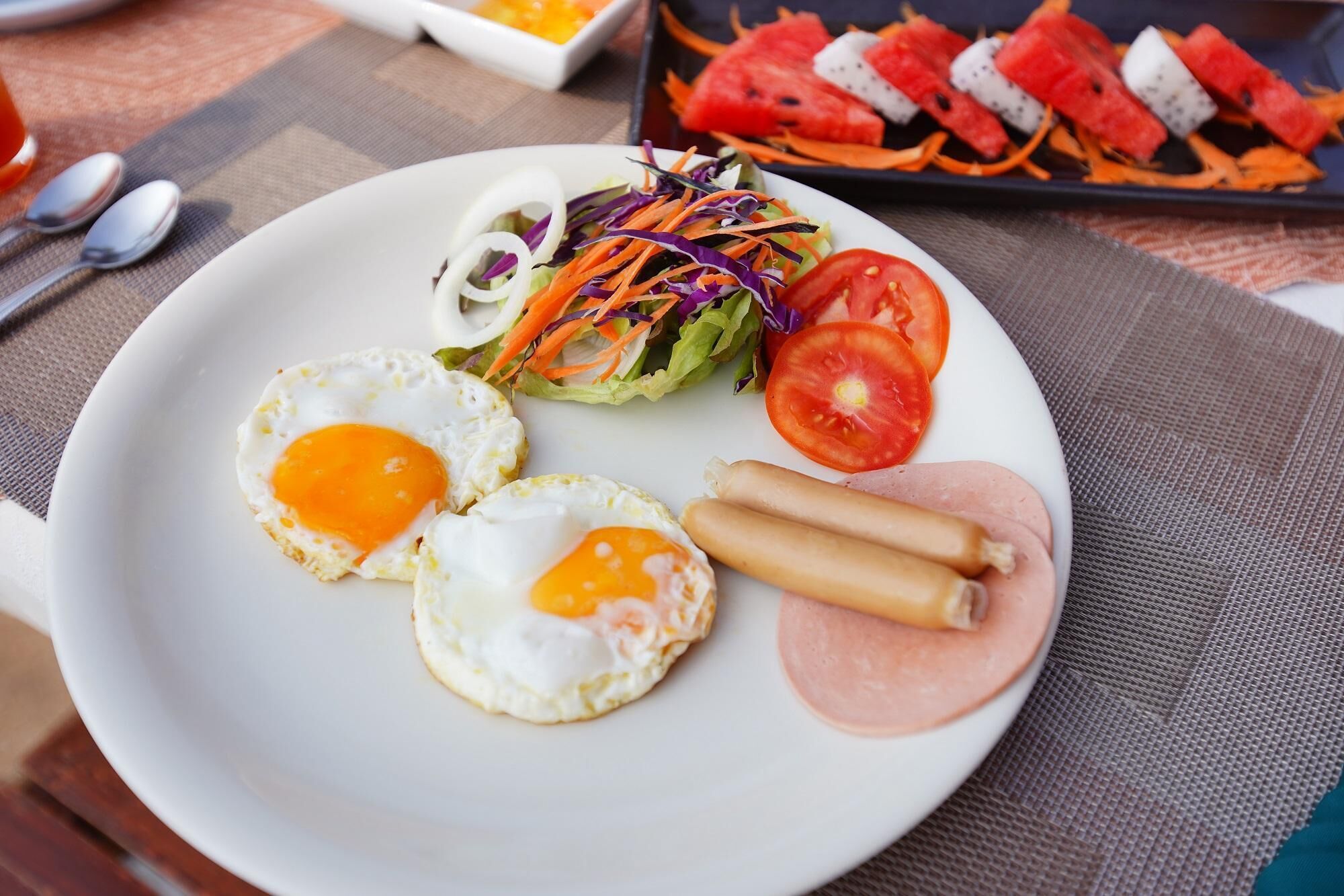 A plate with two sunny-side-up eggs, a slice of tomato, a small salad, and two slices of ham.