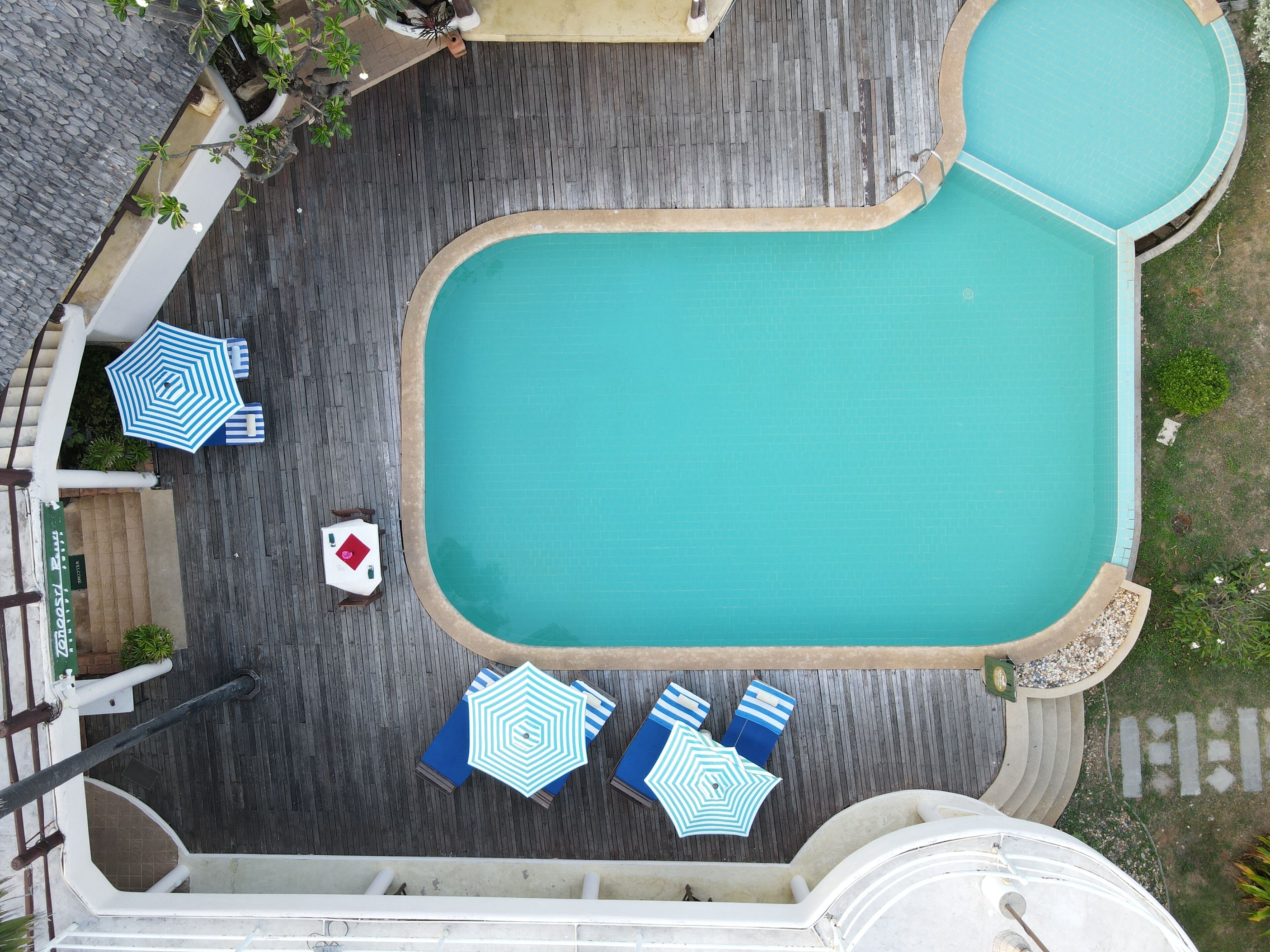 A top-down view of a swimming pool area with a wooden deck, surrounded by lounge chairs and umbrellas.