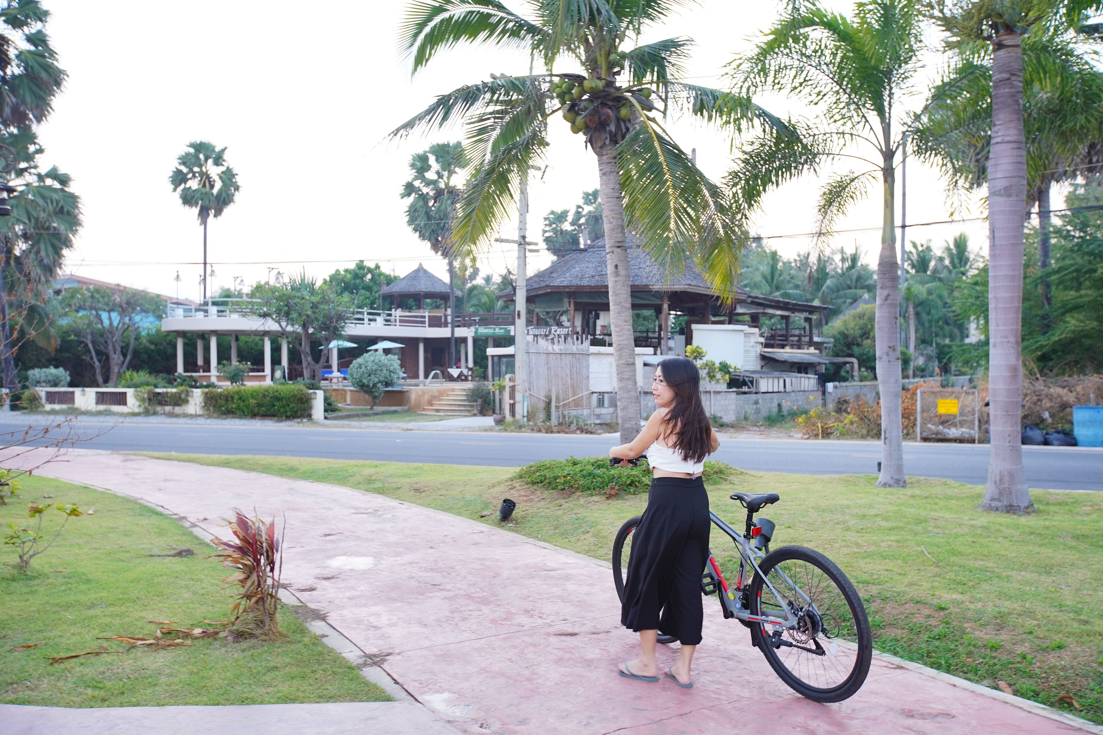 A woman standing next to a bicycle on a sidewalk with palm trees and a building in the background.