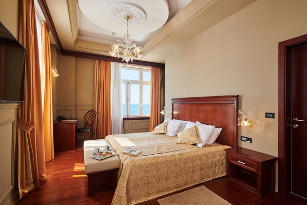 Une chambre d’hôtel avec un grand lit, une table de chevet, un bureau et une chaise. Il y a une fenêtre avec des rideaux, un lustre et un téléviseur fixé au mur.