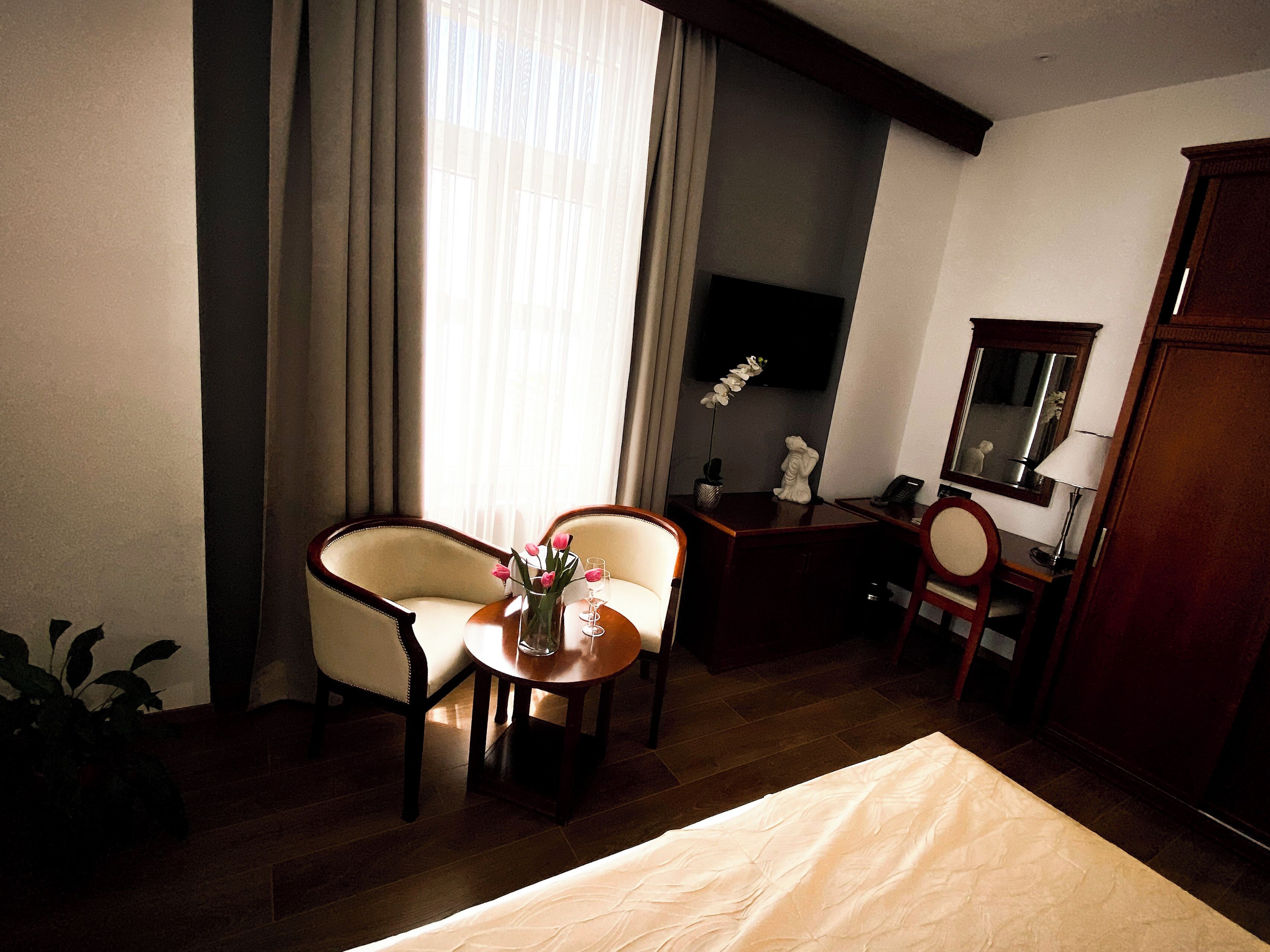 A hotel room with a bed, two armchairs, a small table with a vase of flowers, a desk with a telephone, a mirror, and a television on the wall.