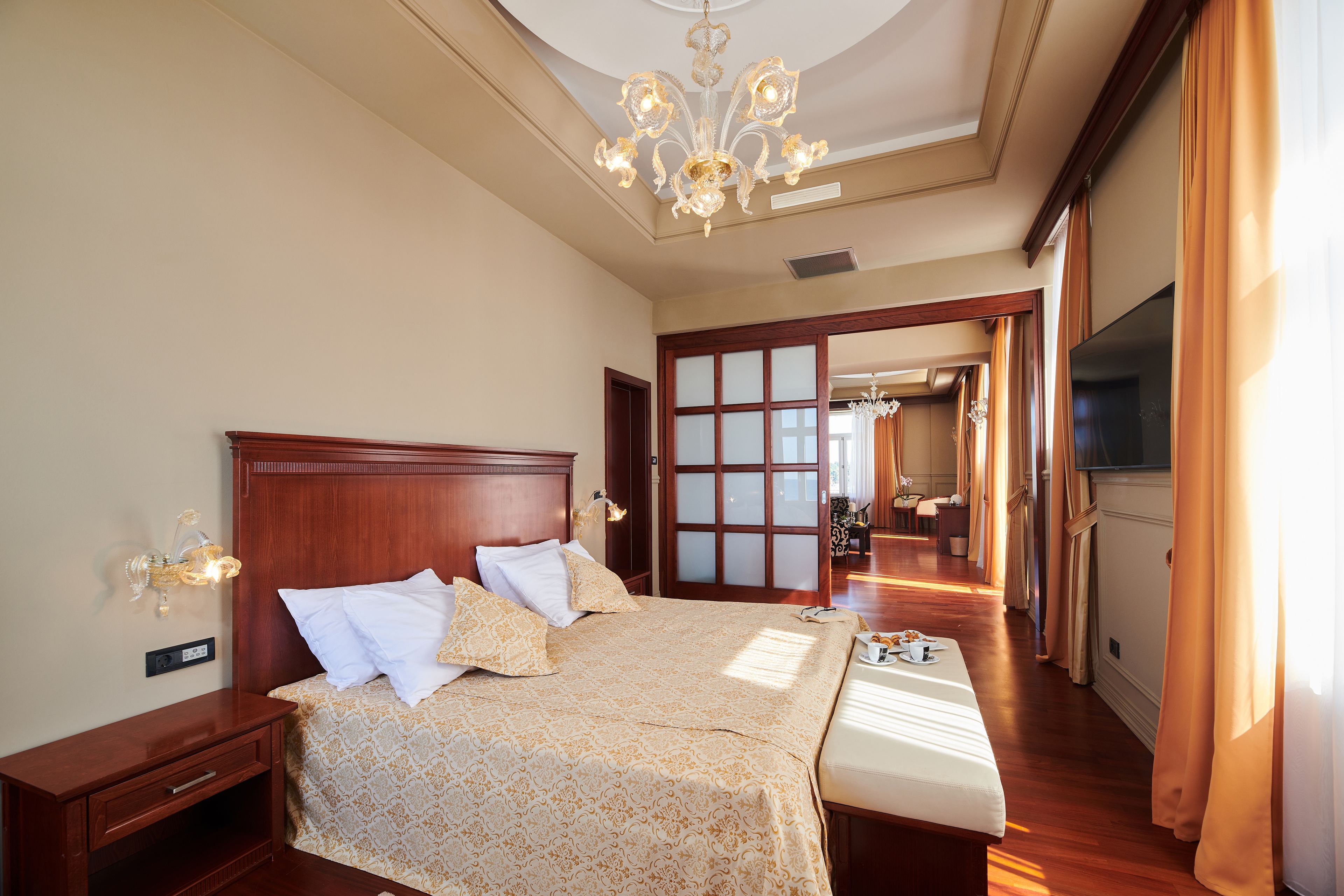 A spacious hotel room with a large bed, a wooden headboard, a chandelier, and a view of the hallway and another room.