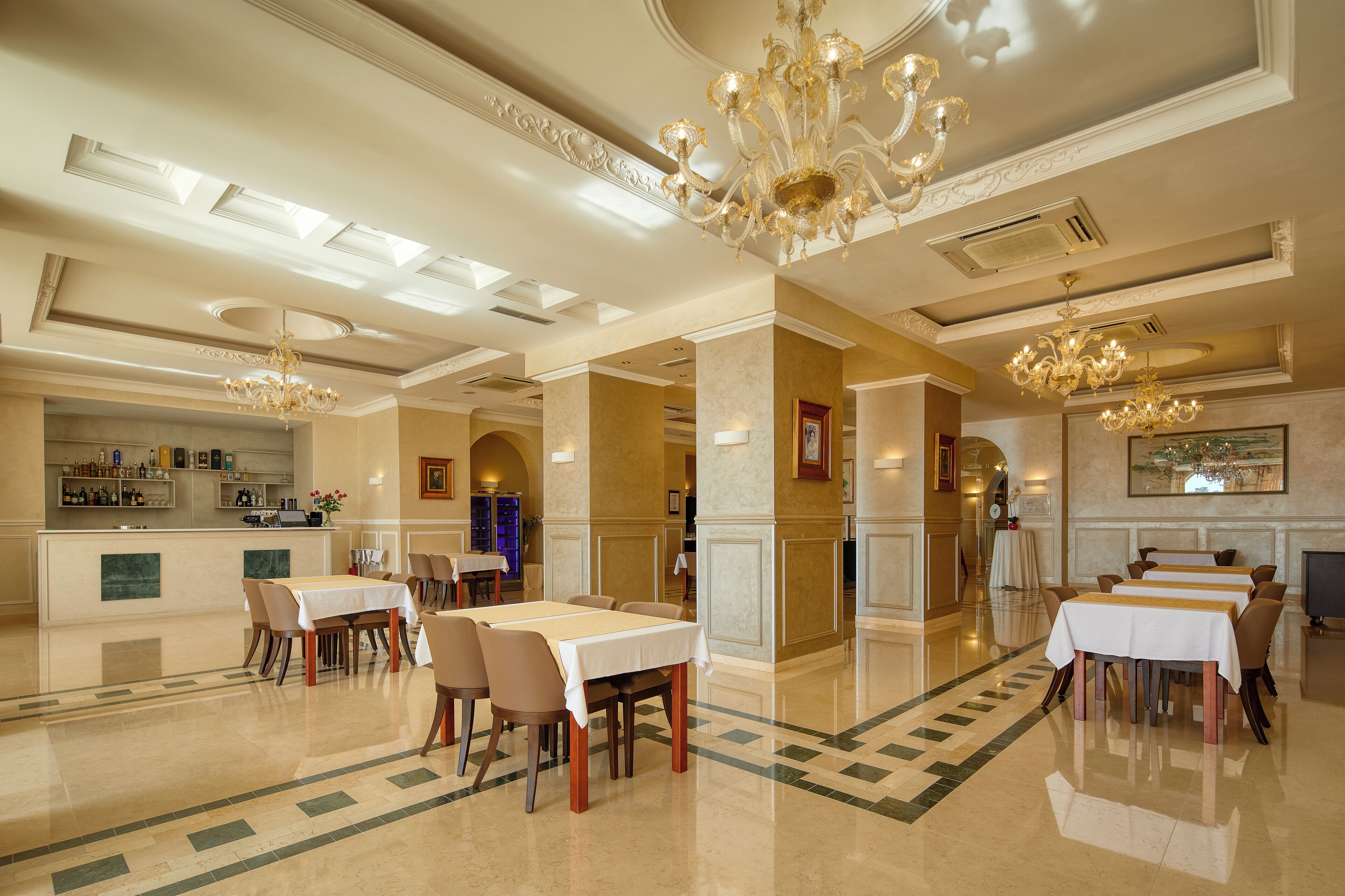 A spacious hotel dining area with multiple tables and chairs, elegant chandeliers, and a well-lit ceiling with skylights.