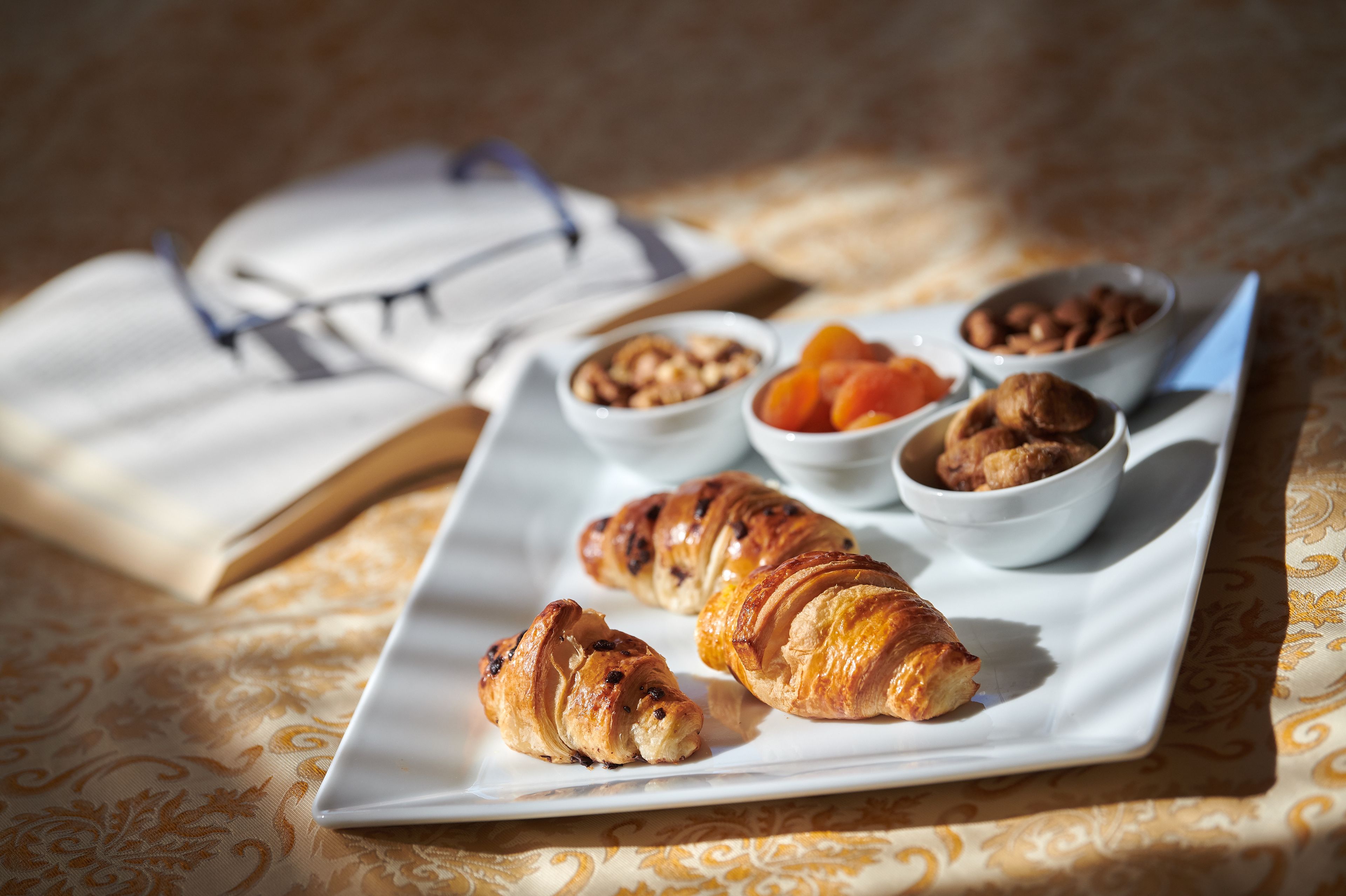 Un plato con cruasanes y tazones de nueces y frutas secas sobre una mesa con un libro y vasos.