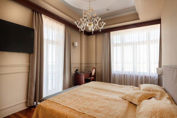 Habitación de hotel con una cama grande, un escritorio con teléfono, una araña y dos ventanas con cortinas.