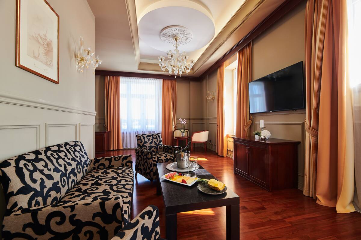 A living room with a sofa, a dining table with a fruit platter, a chandelier, a flat-screen TV, and a large window with curtains.