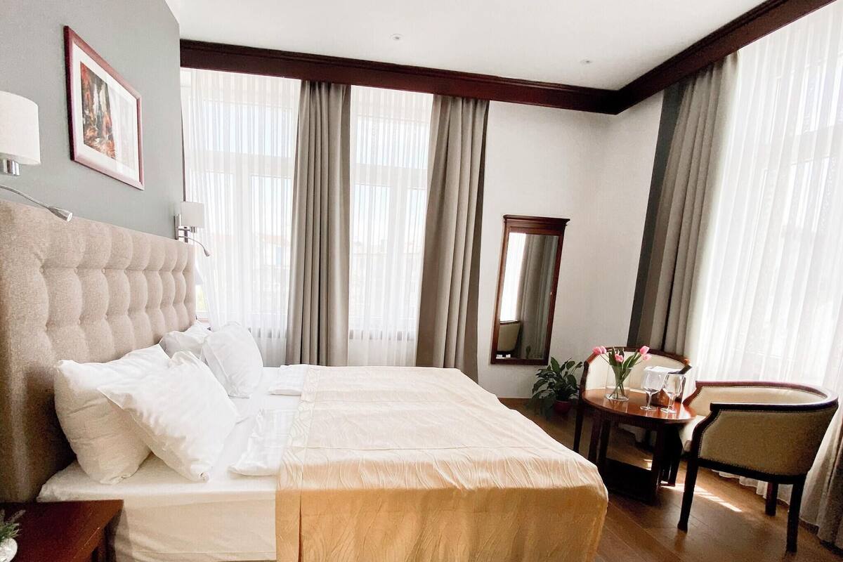 Une chambre d’hôtel avec un grand lit, une table de chevet, une chaise, une petite table avec un vase et des verres, et un miroir.