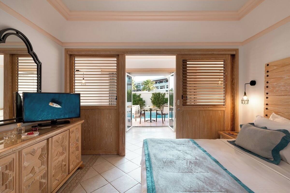 A bedroom with a bed, a TV, wooden cabinets, and a view of an outdoor pool area.