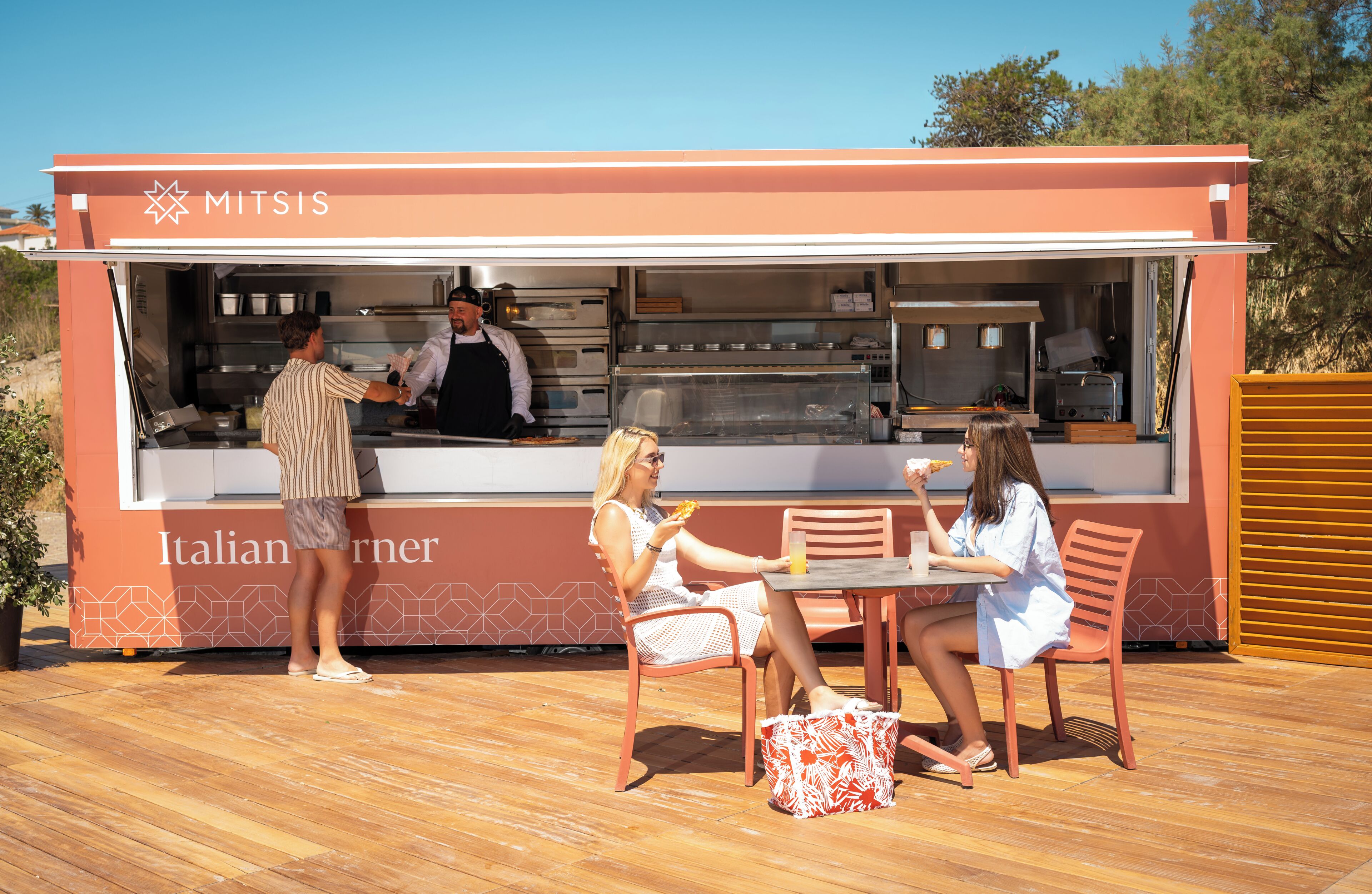 An outdoor food kiosk with a counter, a person serving food, and two customers seated at a table.
