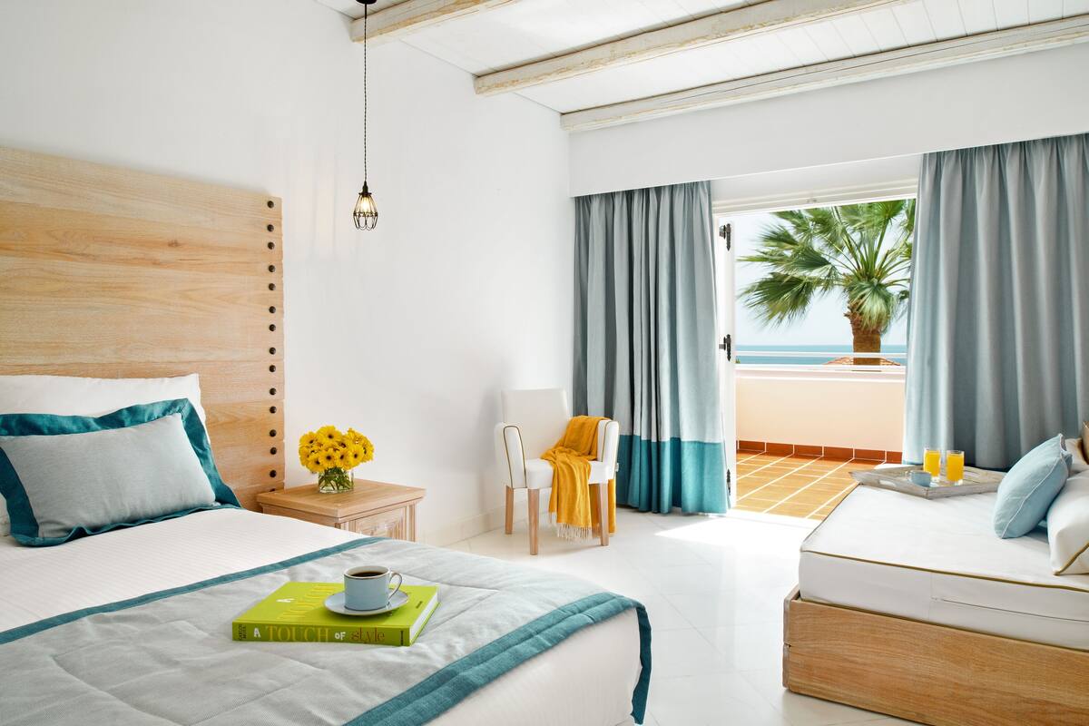 A hotel room with a bed, a side table with a book and a cup, a chair with a yellow throw, and a view of the sea through large windows.