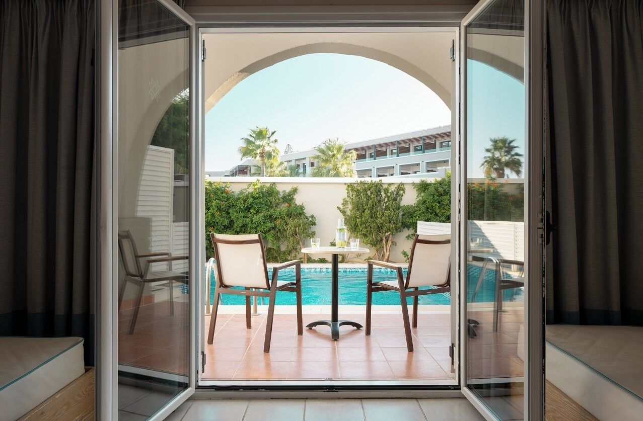 Habitación con puerta de vidrio que da a una zona de piscina, con una mesa y sillas y vistas a la piscina.