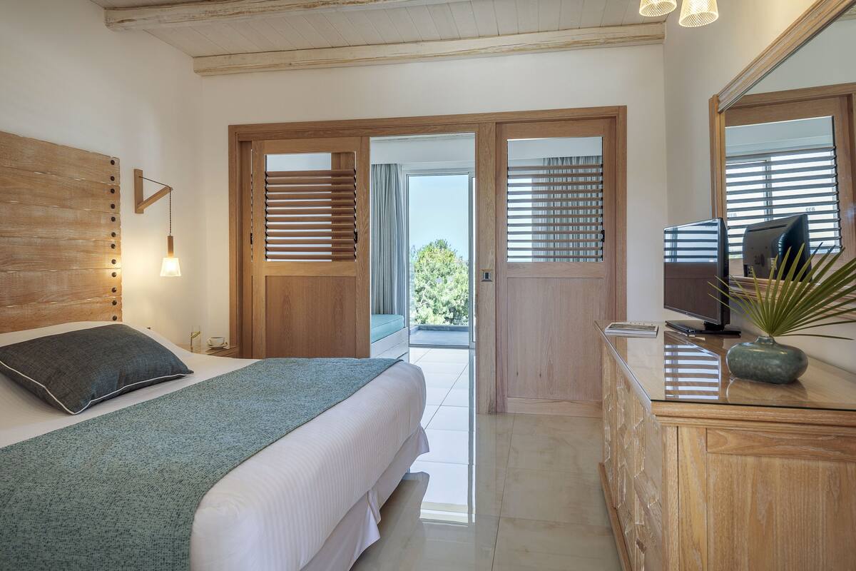 A bedroom with a bed, a dresser, a television, and a view of the outdoors.