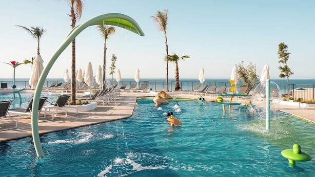 A swimming pool with lounge chairs and umbrellas, palm trees, and a clear sky. - Mitsis Rodos Village (Rhodes)