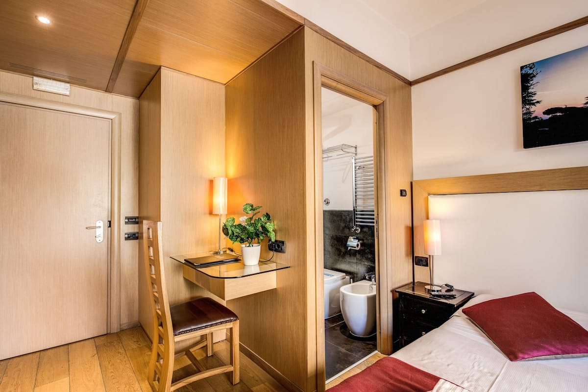 Una camera d'albergo compatta con una scrivania in legno, una sedia, un letto con cuscini rossi, un comodino, una lampada e una pianta in vaso.
