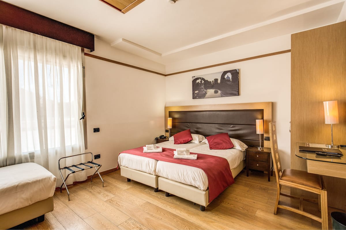 Una camera d'albergo con un letto, una scrivania, una sedia, un comodino e una finestra con tende.
