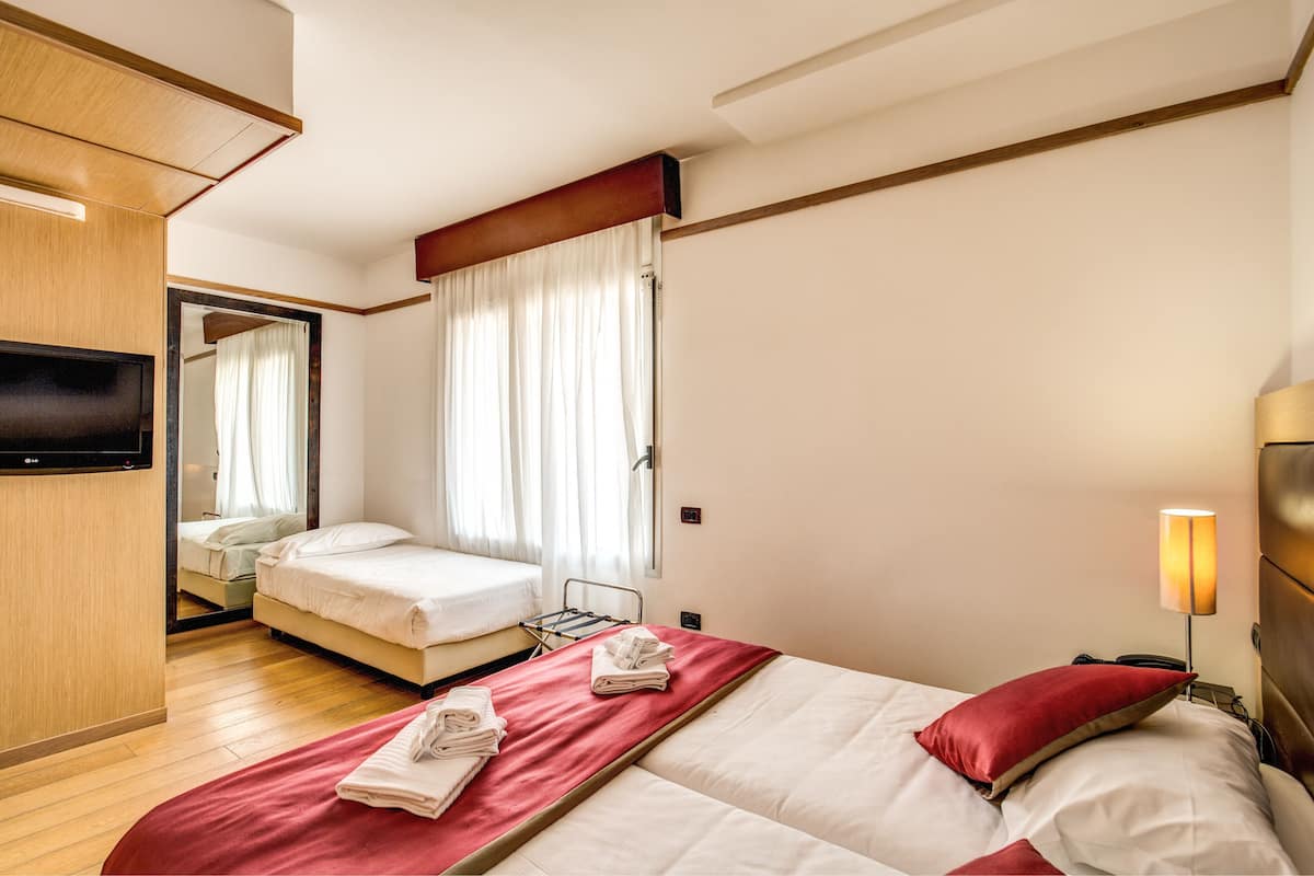 Una camera d'albergo con due letti, una televisione, un comodino, una lampada e una finestra con tende.