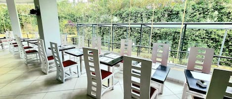 A restaurant with white chairs and tables, large windows, and a view of greenery outside.
