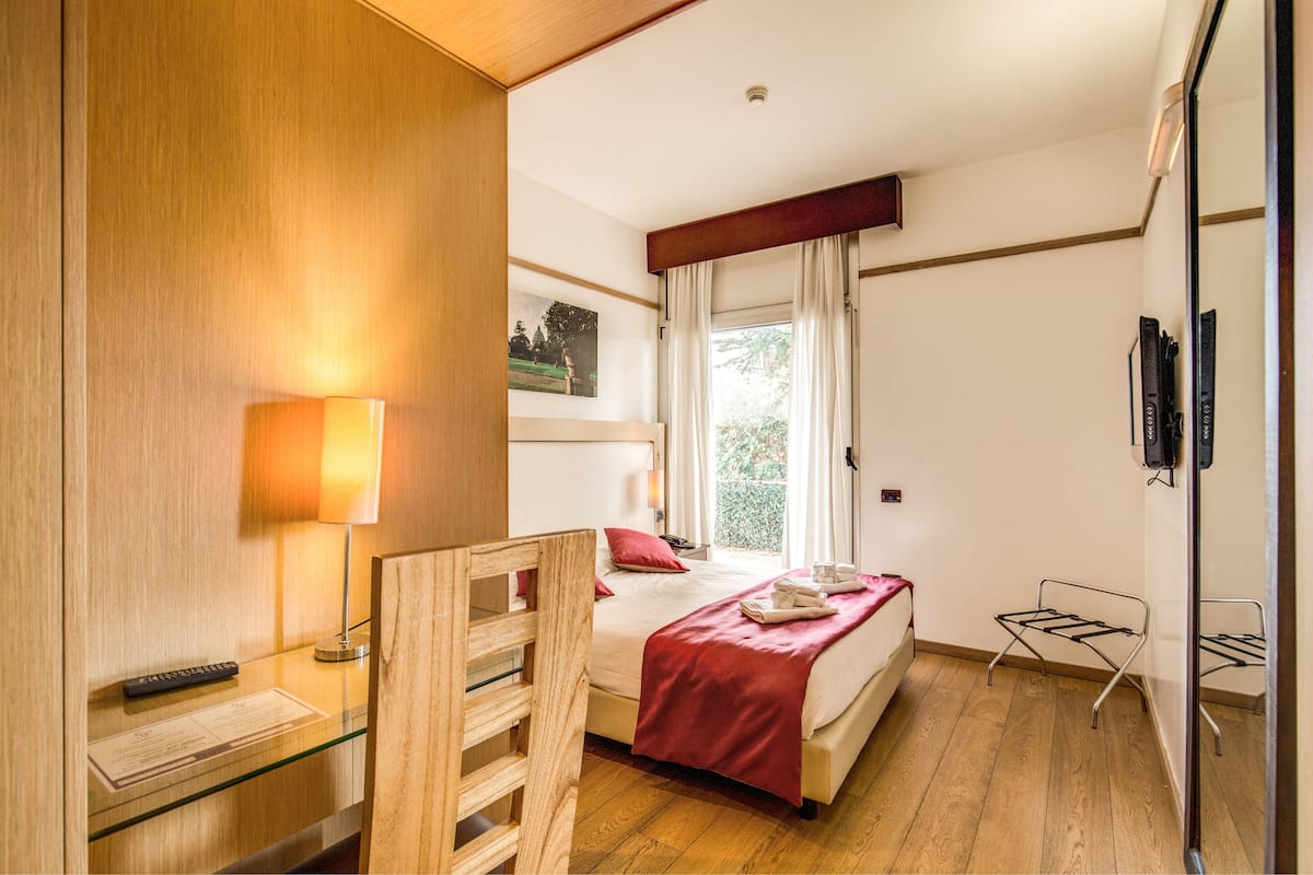 Una camera d'albergo con una scrivania in legno, un letto con coperta rossa, una finestra con tende e uno specchio a parete.