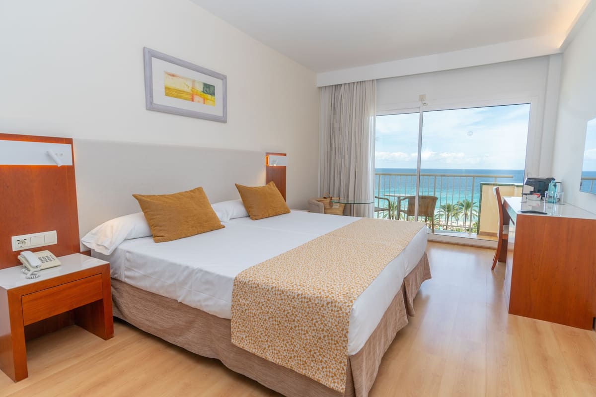 Ein Hotelzimmer mit Bett, Schreibtisch, Stuhl, Telefon, Fenster mit Meerblick und einem Bild an der Wand.