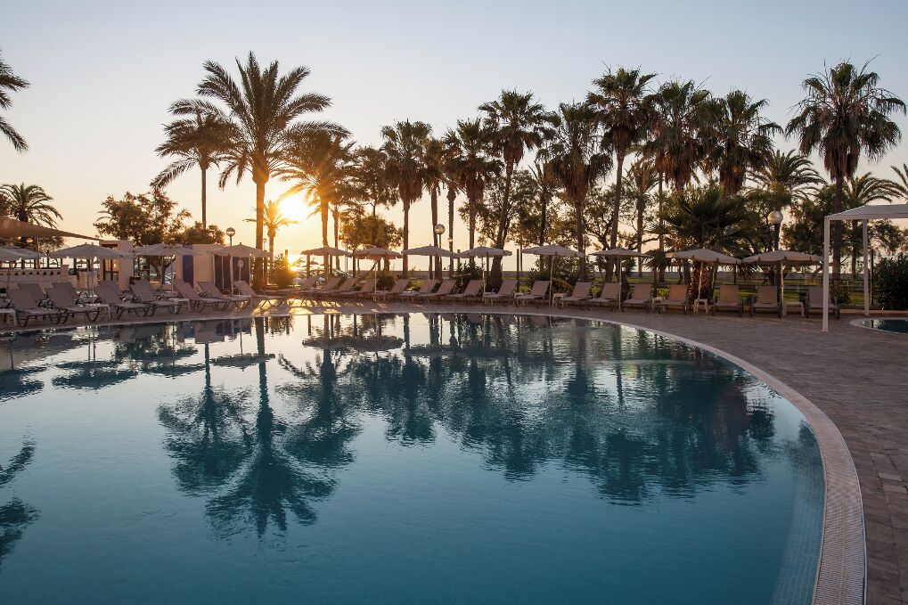 Una piscina con sillas de descanso y sombrillas, rodeada de palmeras.