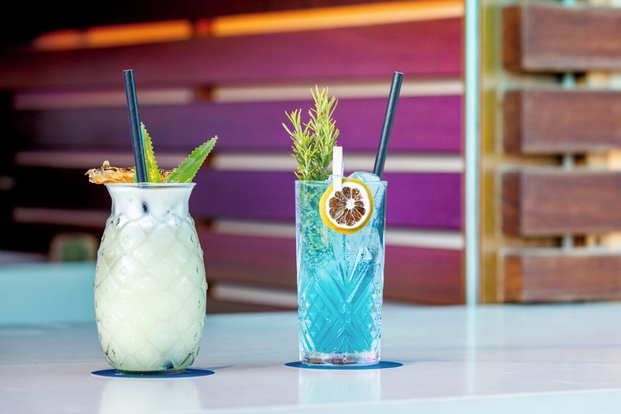 Two drinks in glass containers with straws, one with a pineapple garnish and the other with a lemon slice and mint.