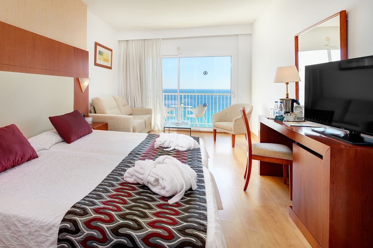 Ein Hotelzimmer mit einem großen Bett, einem Schreibtisch mit Fernseher, einem Sessel und einem Balkon mit Meerblick.